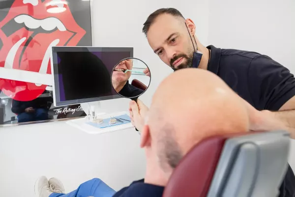 Zahnarzt hält einen Spiegel, in dem der Patient seine Zähne betrachtet und eine Zahnbürste benutzt. Im Hintergrund ein Rolling-Stones-Poster.